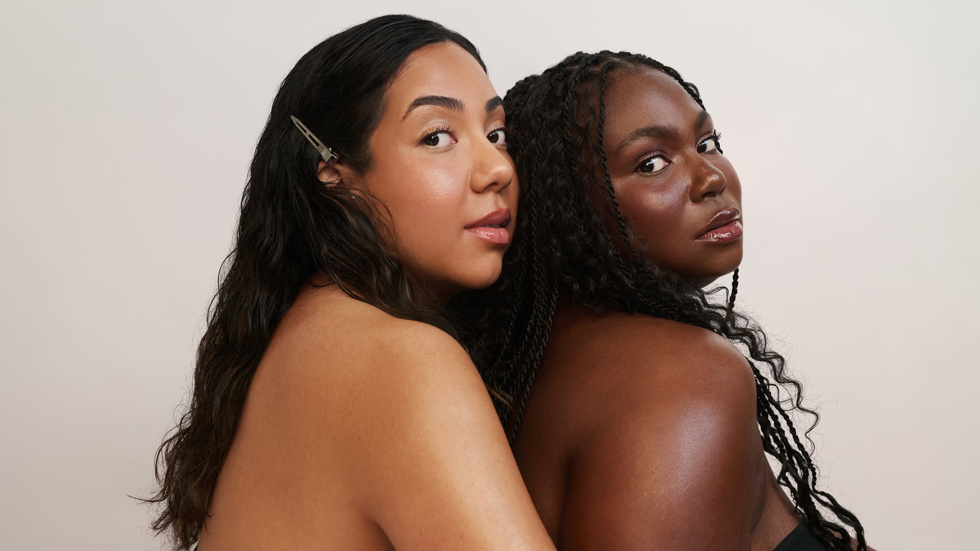 Deux femmes posent rapprochées l'une de l'autre, regardant la caméra par-dessus leur épaule. L'une a de longs cheveux foncés ondulés avec une barrette ; l'autre a de longs cheveux tressés. Toutes deux portent des hauts noirs sans bretelles.