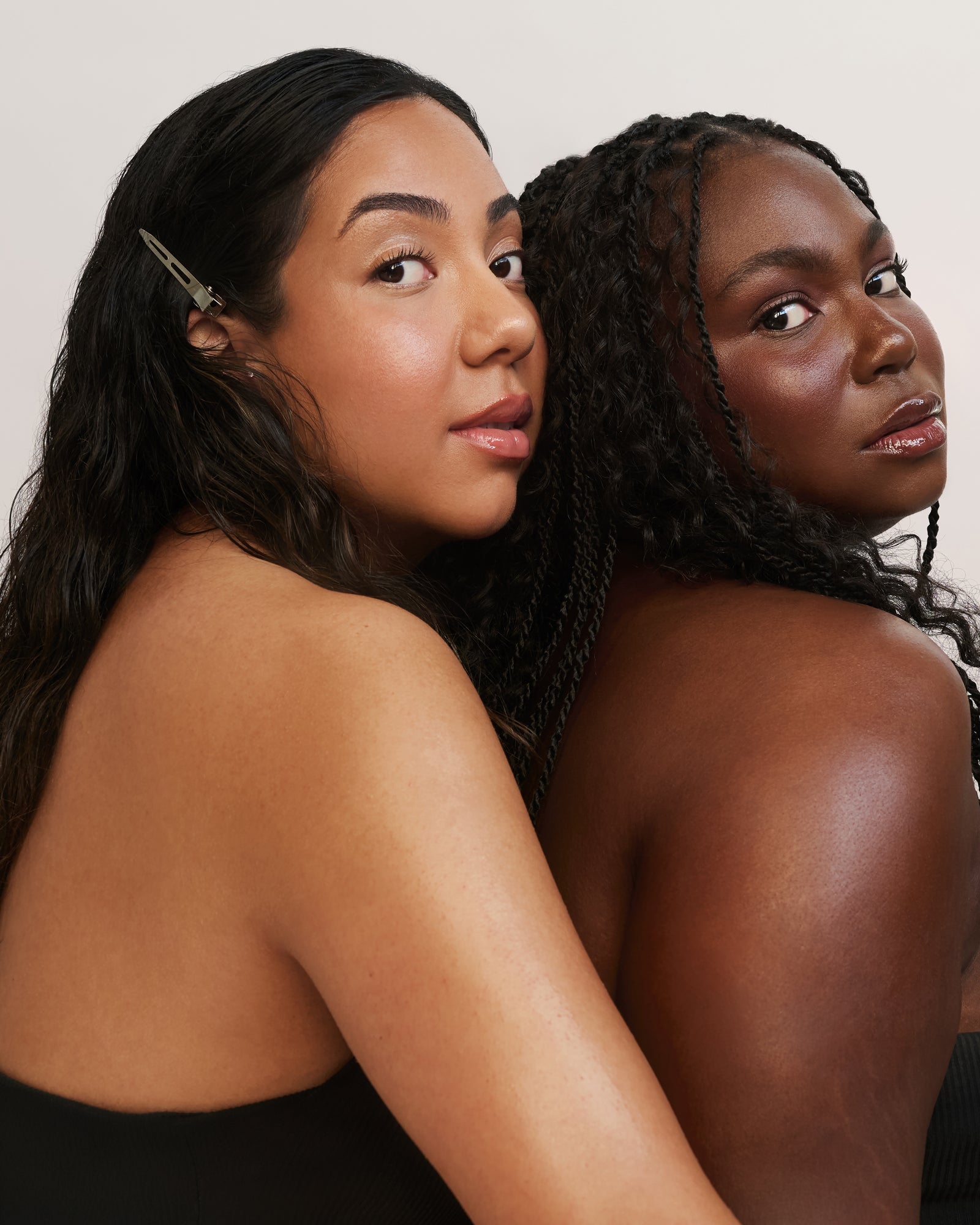 Deux femmes posent rapprochées l'une de l'autre, regardant la caméra par-dessus leur épaule. L'une a de longs cheveux foncés ondulés avec une barrette ; l'autre a de longs cheveux tressés. Toutes deux portent des hauts noirs sans bretelles.