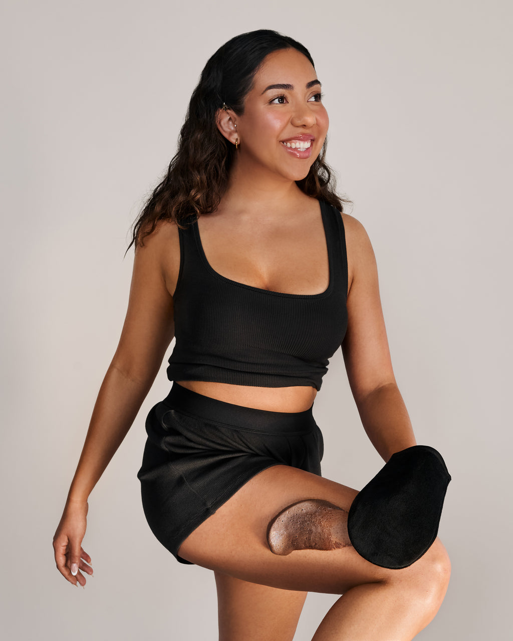 Une femme hispanique aux cheveux noirs pose en mouvement, vêtue d'un ensemble noir. Elle applique la mousse autobronzante Ultra foncé de TravelMe avec un gant applicateur, sur sa jambe.