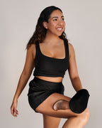 Une femme hispanique aux cheveux noirs pose en mouvement, vêtue d'un ensemble noir. Elle applique la mousse autobronzante Ultra foncé de TravelMe avec un gant applicateur, sur sa jambe.