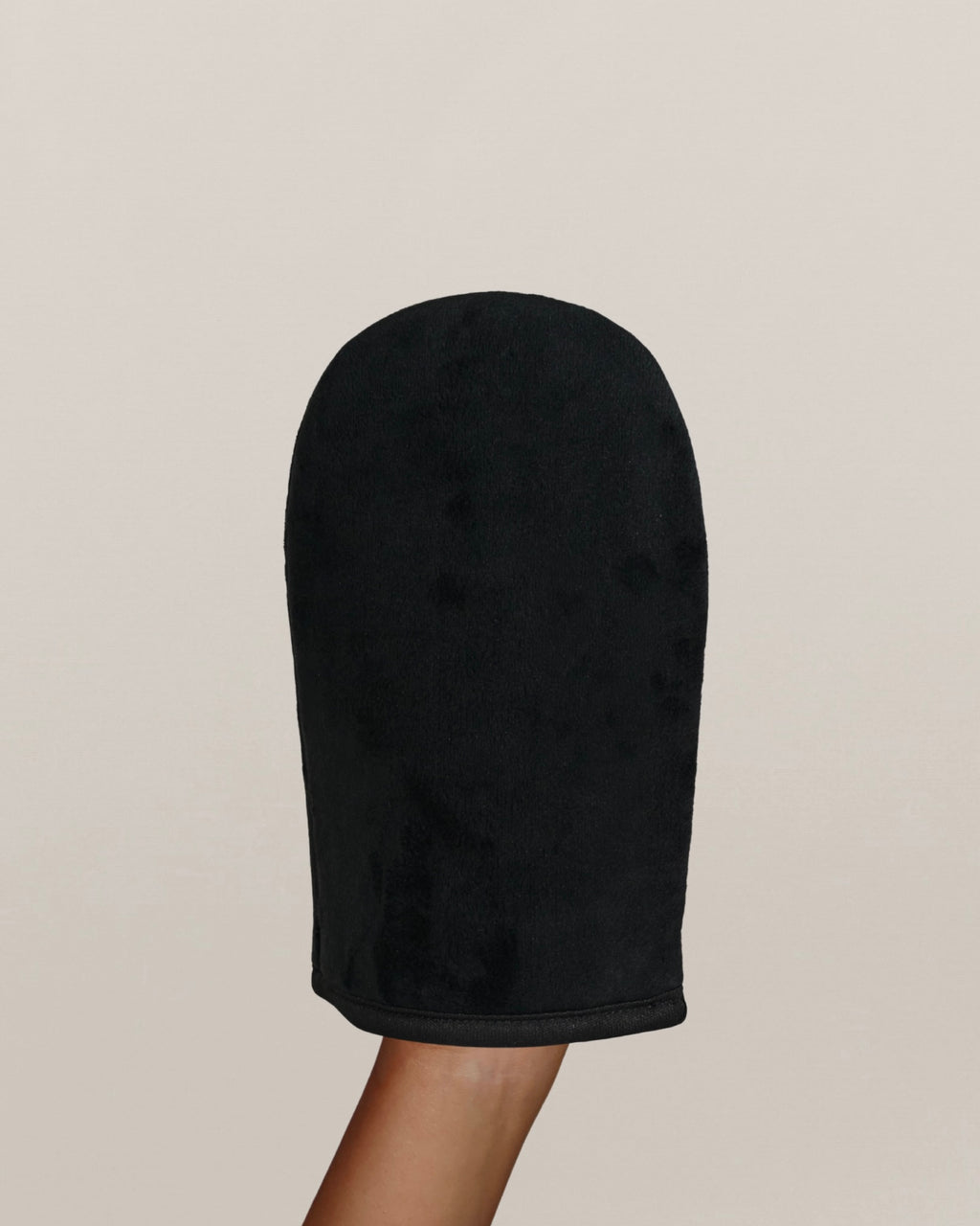 Une main porte un gant d'application d'autobronzant noir sur un fond uni beige clair.