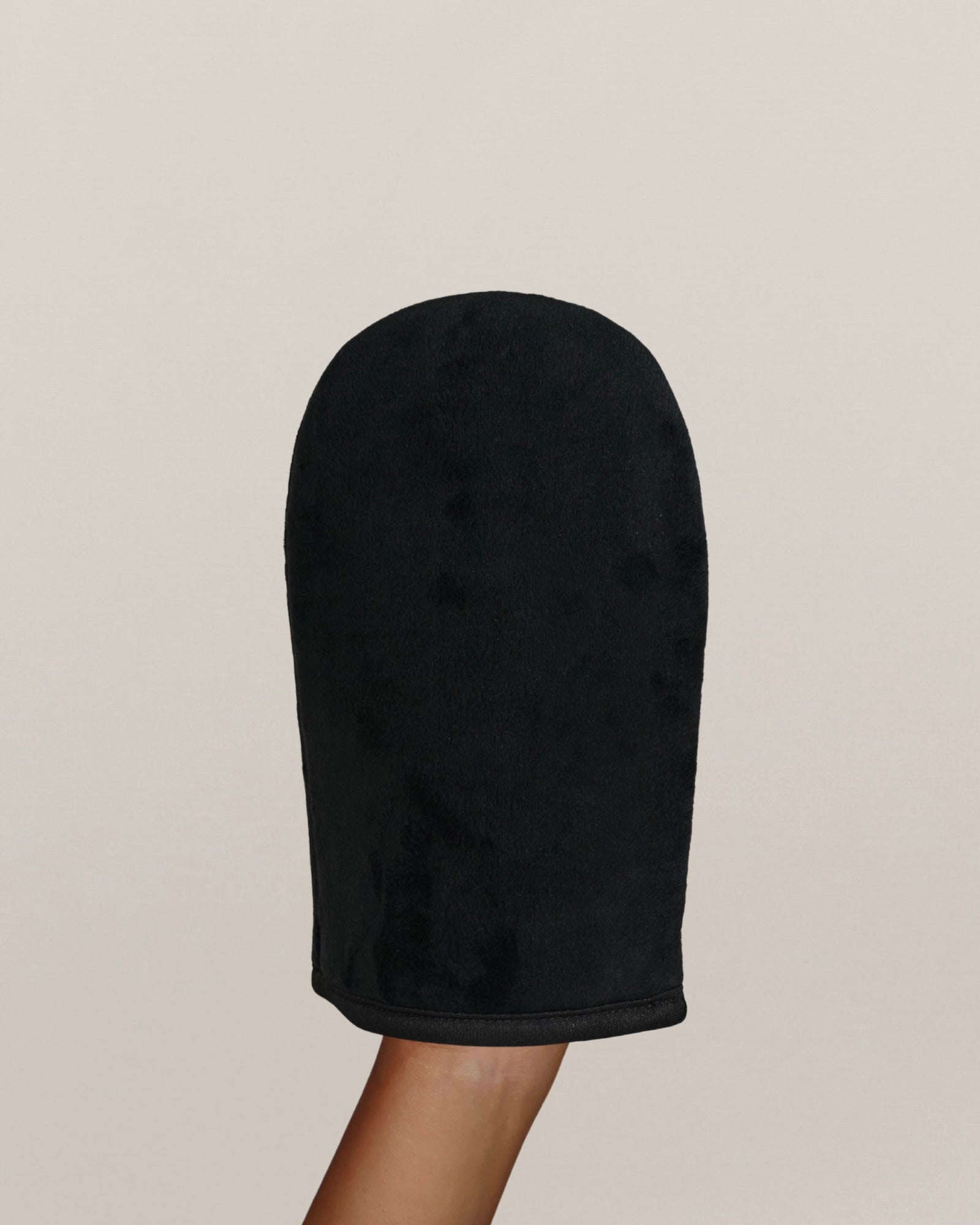 Une main porte un gant d'application d'autobronzant noir sur un fond uni beige clair.