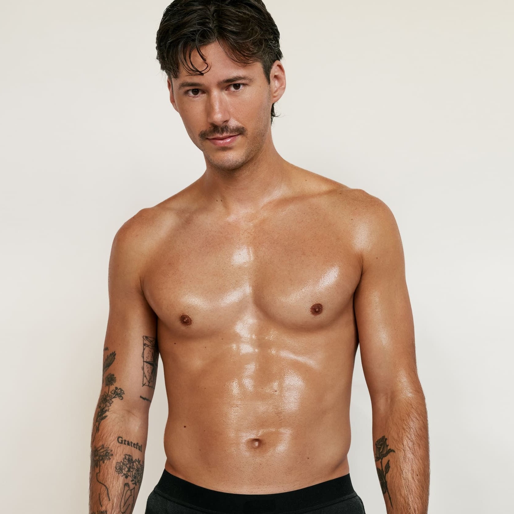Un homme torse nu, tatoué et moustachu, vêtu d'un short noir, se tient sur un fond uni