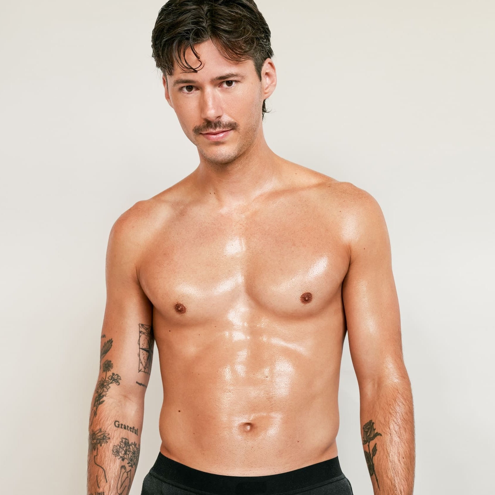 Un homme torse nu, tatoué et moustachu, vêtu d'un short noir, se tient sur un fond uni