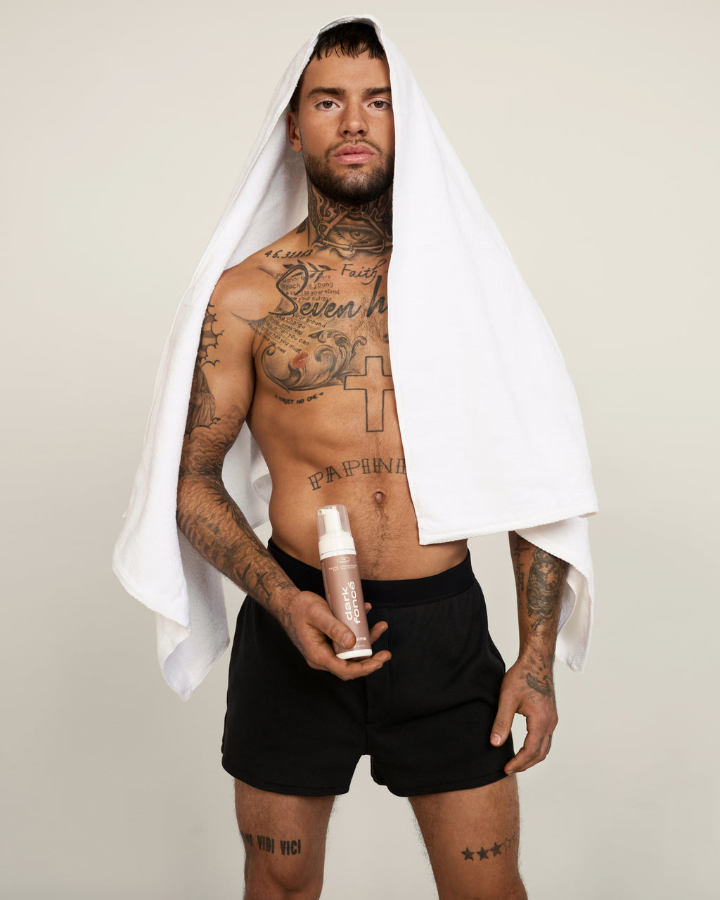 Un homme torse nu, tatoué et barbu, vêtu d'un short noir, a une serviette blanche sur la tête et se tient sur un fond uni. Il tient dans sa main un e mousse autobronzante de la teinte Foncé - un produit autobronzant à ajouter à sa routine bien-être
