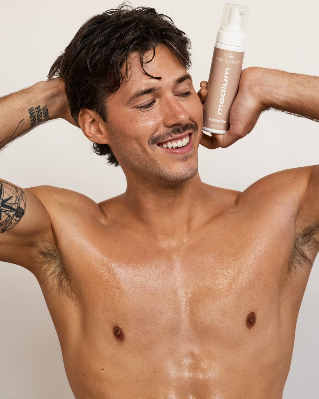 Un homme torse nu, tatoué et moustachu, sourit avec les mains de chaque côté de sa tête et tient dans ses mains une mousse autobronzante Medium de TravelMe