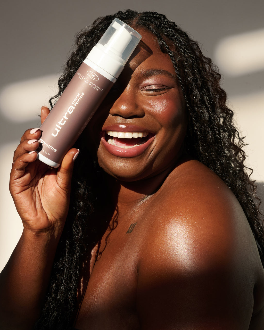 Une femme aux longs cheveux tressés tient une mousse autobronzante ultra foncé de TravelMe devant son visage, souriant à la caméra.