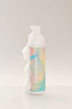 Produit Effaceur d'autobronzant à l'emballage multicolore et métallique, avec de la mousse blanche qui coule, de l'entreprise québécoise TravelMe, sur un fond blanc.