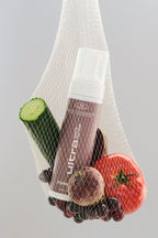 Un sac en filet blanc contenant un flacon de la mousse autobronzante Ultra foncé, ainsi qu'un concombre, une tomate et plusieurs fruits, sur un fond gris neutre.