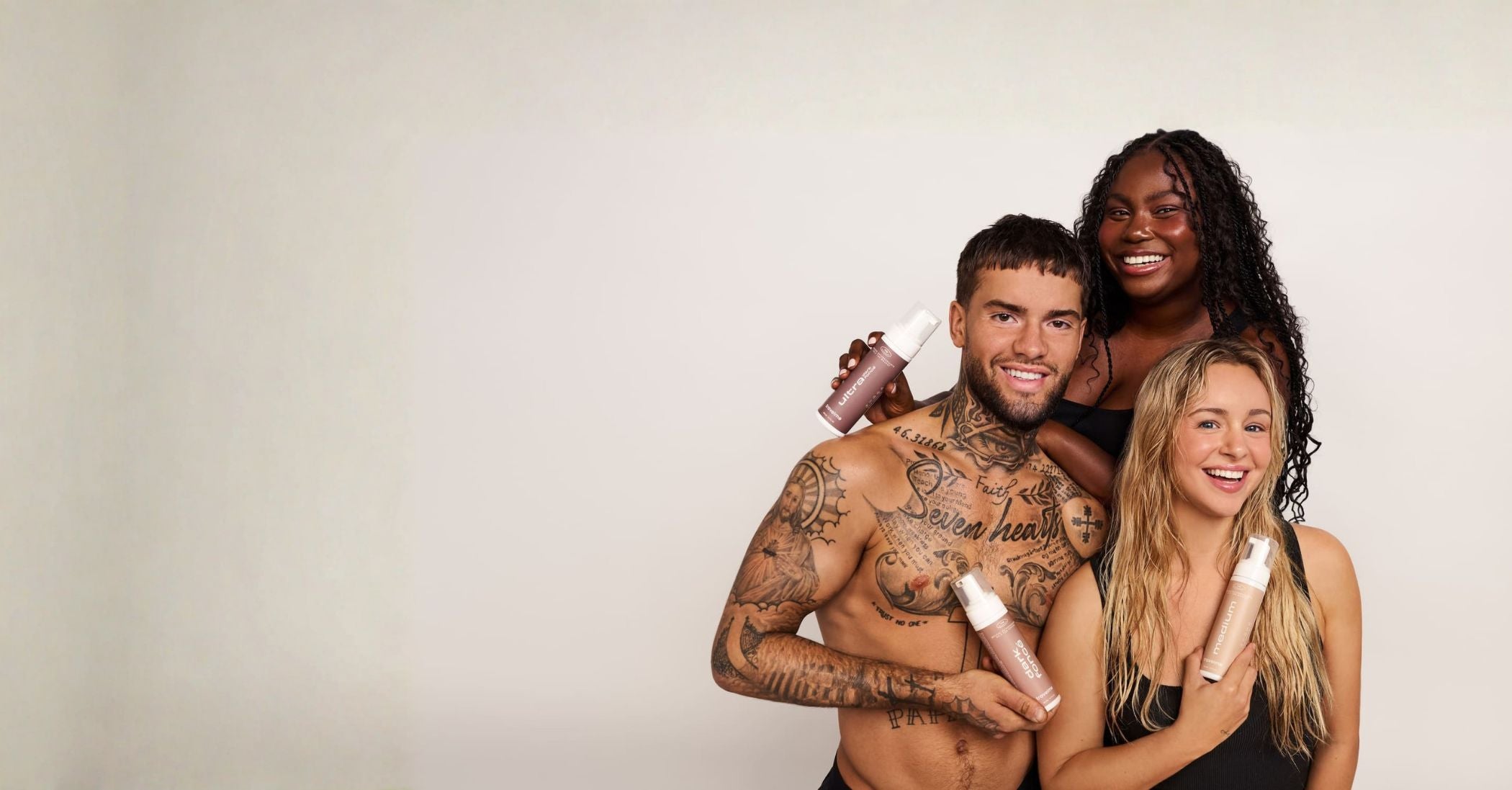Trois personnes souriantes, deux femmes et un homme, posant ensemble sur un fond uni, chacun tenant un flacon de mousse autobronzante de la marque québécoise TravelMe. Produit autobronzant à ajouter naturellement à sa routine bien-être